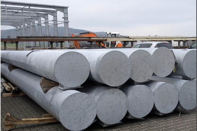 Lagerfläche mit mehreren großen, zylindrischen Stahlrohren auf Holzpaletten, im Hintergrund leichte Industriearchitektur.