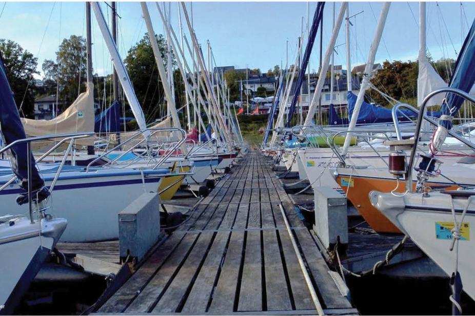 Holzsteg zwischen mehreren Segelbooten im Hafen, umgeben von Bäumen und Gebäuden im Hintergrund.