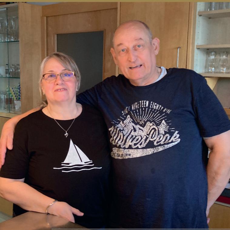 Paar in einer Küche, stehen nahe beieinander, lächeln in die Kamera, tragen lässige Freizeitkleidung.