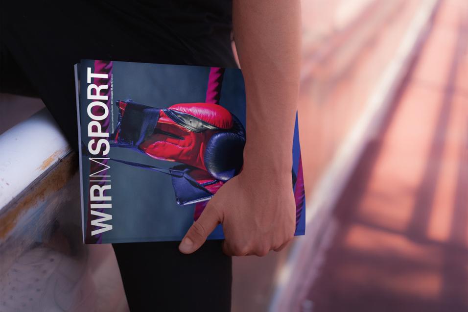 Hand hält ein Magazin mit dem Titel „WIR IM SPORT“ auf einer Sportbahn.